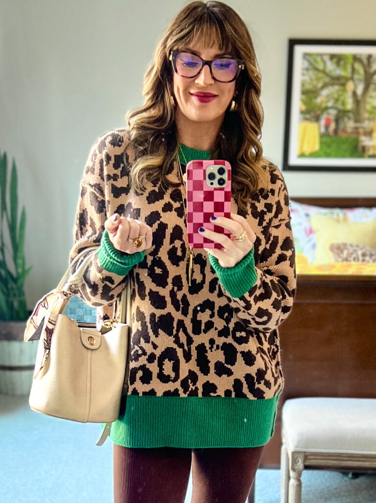 Mirror selfie of woman carrying beige Amazon mini bucket handbag with silk scarf on handle, wearing leopard print sweater with green trim, burgundy pants, tortoise glasses, layered gold necklaces, pink checkered phone case, colorful bedroom visible in background