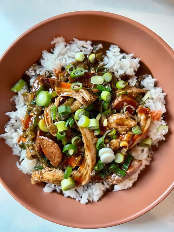 Overhead view of chicken and sausage gumbo served over white rice in a terracotta bowl, topped with sliced scallions and hot sauce