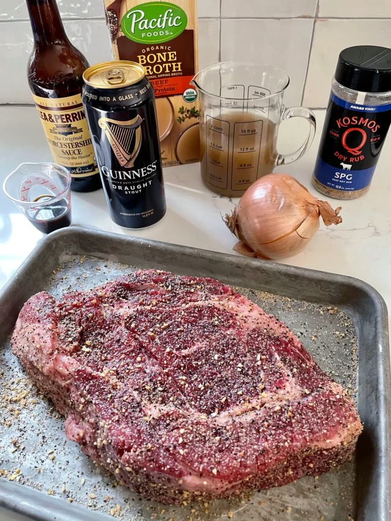 Overhead view of French Dip ingredients: seasoned chuck roast on a sheet pan with Guinness, Worcestershire sauce, beef broth, onion, and dry rub