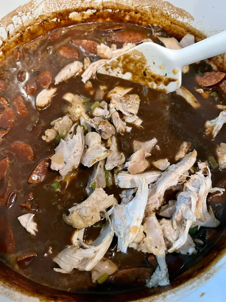 Adding shredded chicken back into the pot of gumbo with andouille sausage
