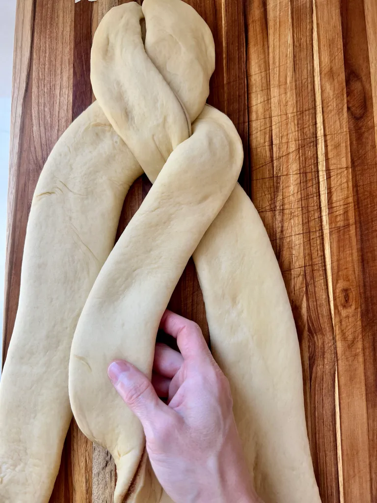 Hands braiding the three strips of cinnamon-filled dough on a wooden surface