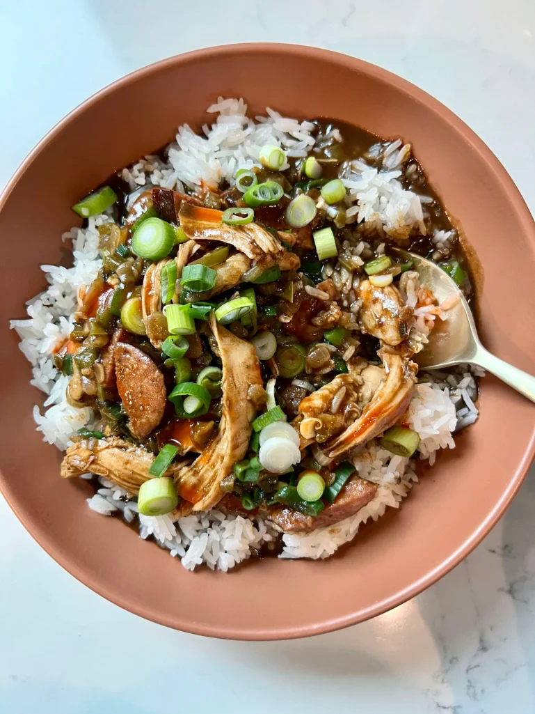 Overhead view of a bowl of chicken and sausage gumbo over rice with a spoon, topped with scallions and hot sauce