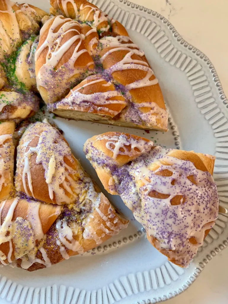 Sliced King Cake on a platter showing a piece pulled away, decorated with purple, green, and gold sugars