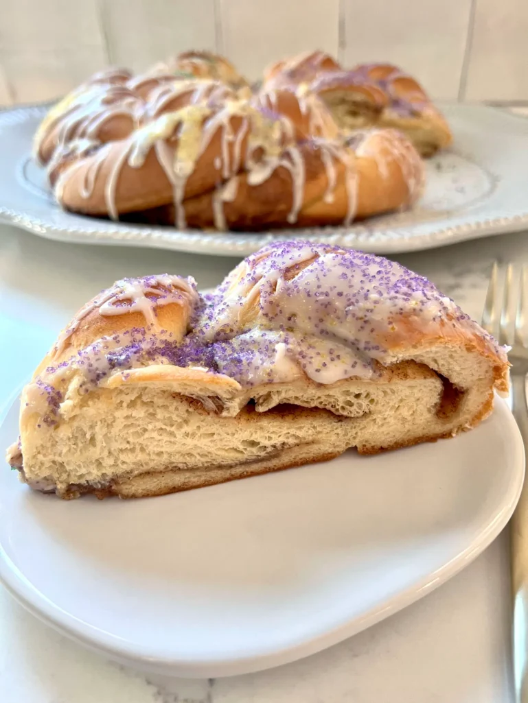 Slice of King Cake on a small plate showing the soft brioche interior and cinnamon sugar swirl, with the whole cake in the background