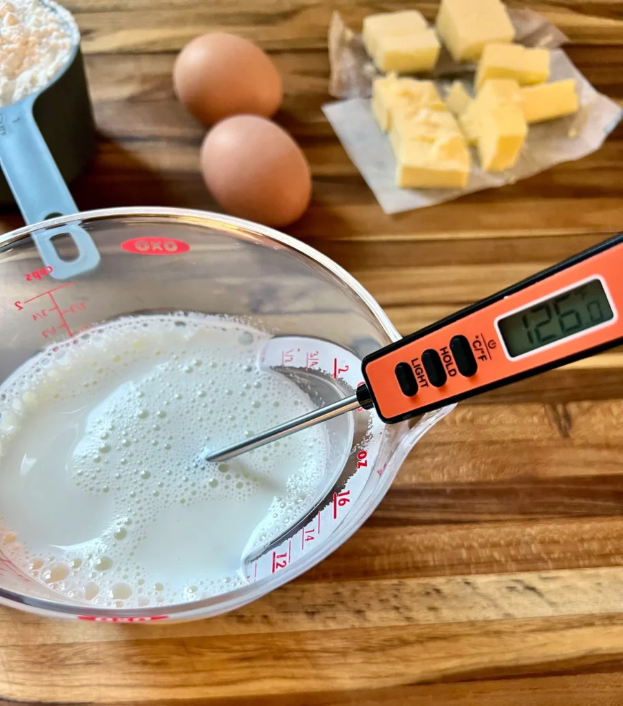 Checking the temperature of warmed milk with a digital thermometer reading 126°F, with eggs and butter in the background