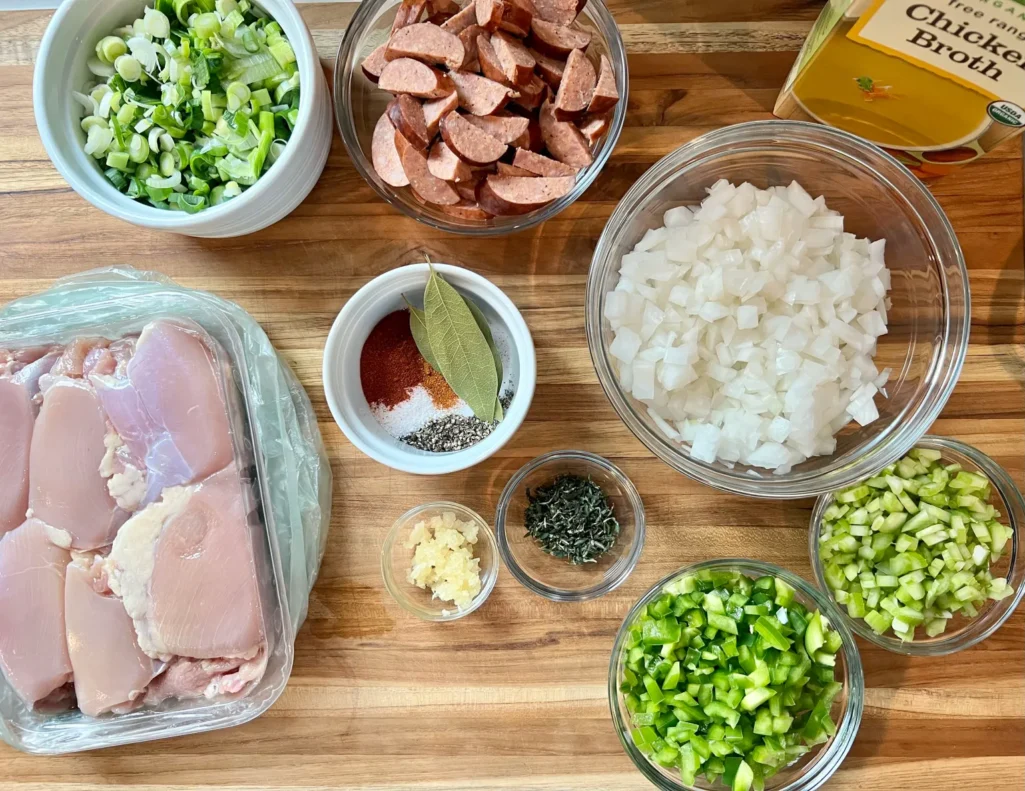 Overhead view of gumbo ingredients: chicken thighs, andouille sausage, onion, celery, bell pepper, scallions, garlic, thyme, spices, and chicken broth