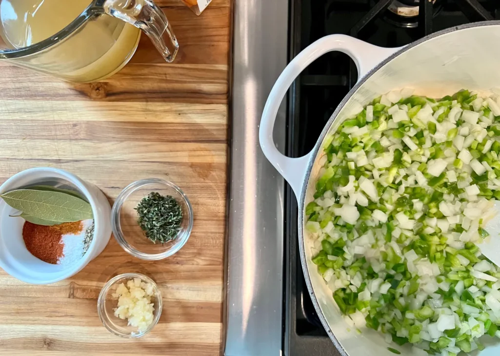 Holy trinity of onion, celery, and bell pepper sautéing in a Dutch oven with seasonings and broth nearby