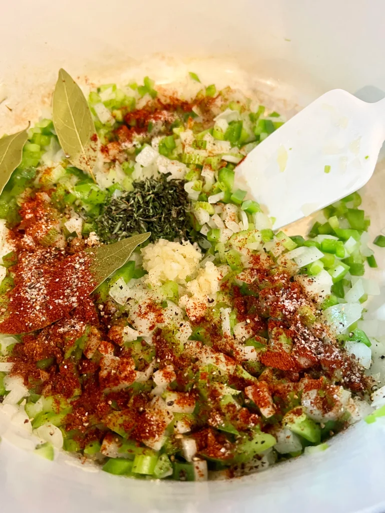 Adding garlic, thyme, paprika, cayenne, bay leaves, and black pepper to the sautéed vegetables