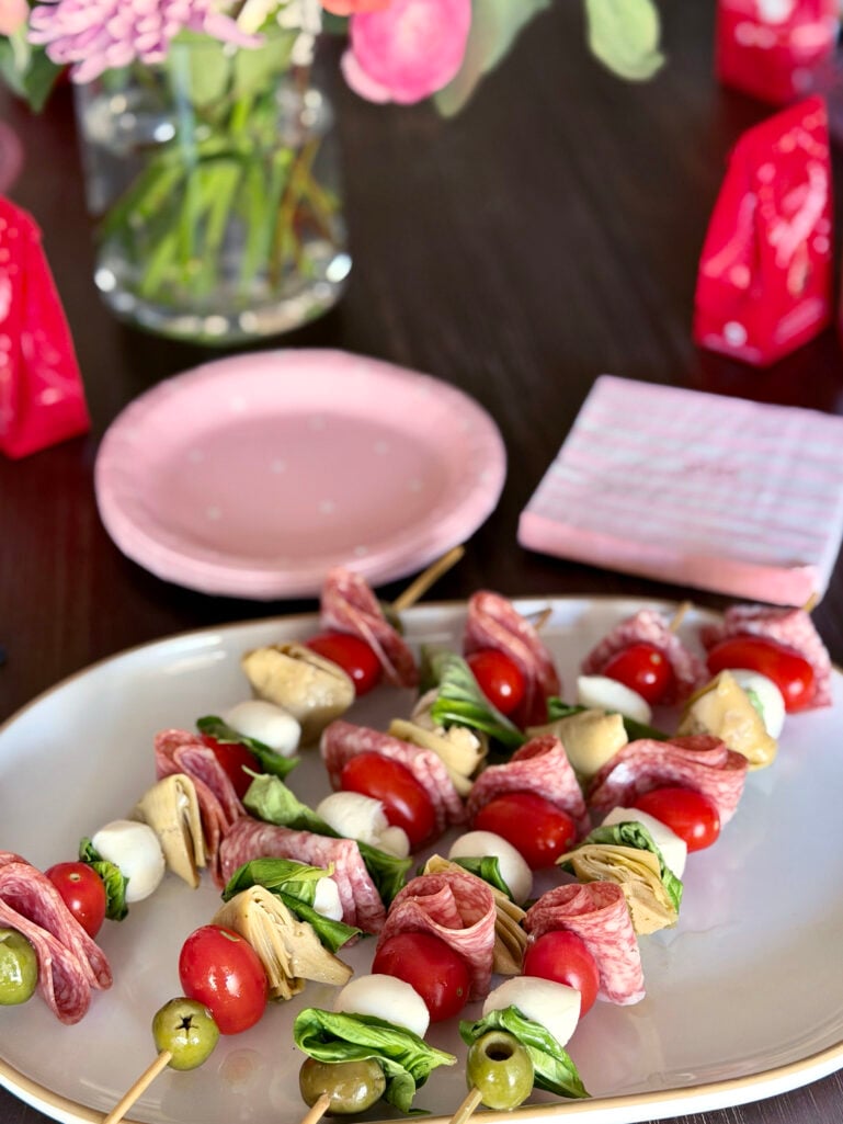 Antipasti skewers with salami, fresh mozzarella, cherry tomatoes, marinated artichoke hearts, green olives, and basil on a white platter, with pink polka-dot plates and Valentine's flowers in the background
