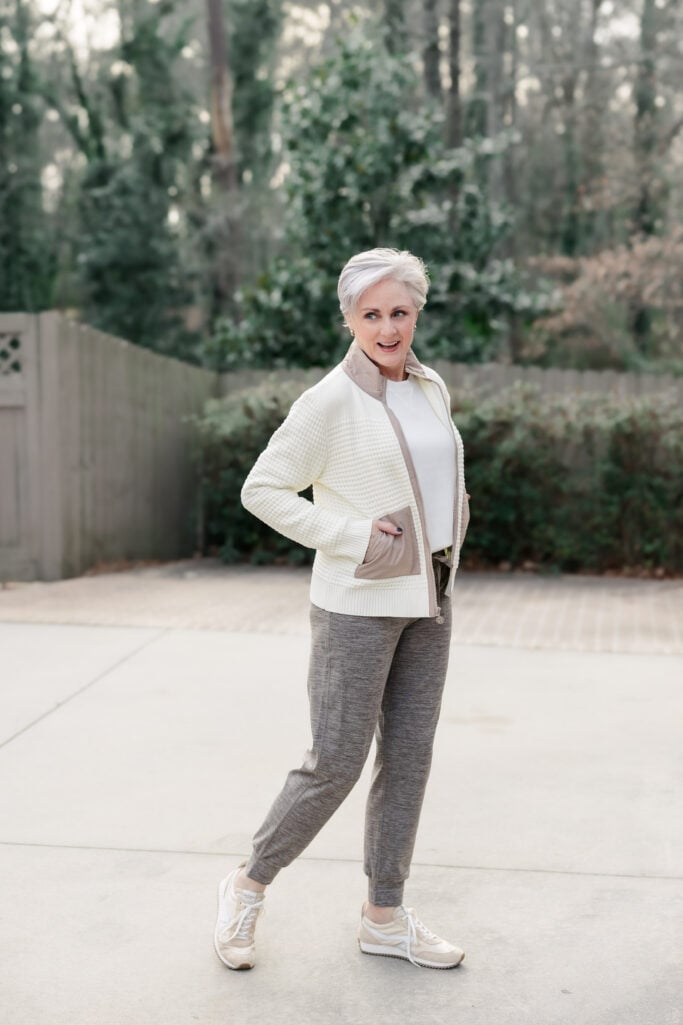 Woman over 50 with silver hair wearing cream waffle-knit sweater jacket with taupe trim over white tee and heather brown drawstring joggers with neutral retro sneakers