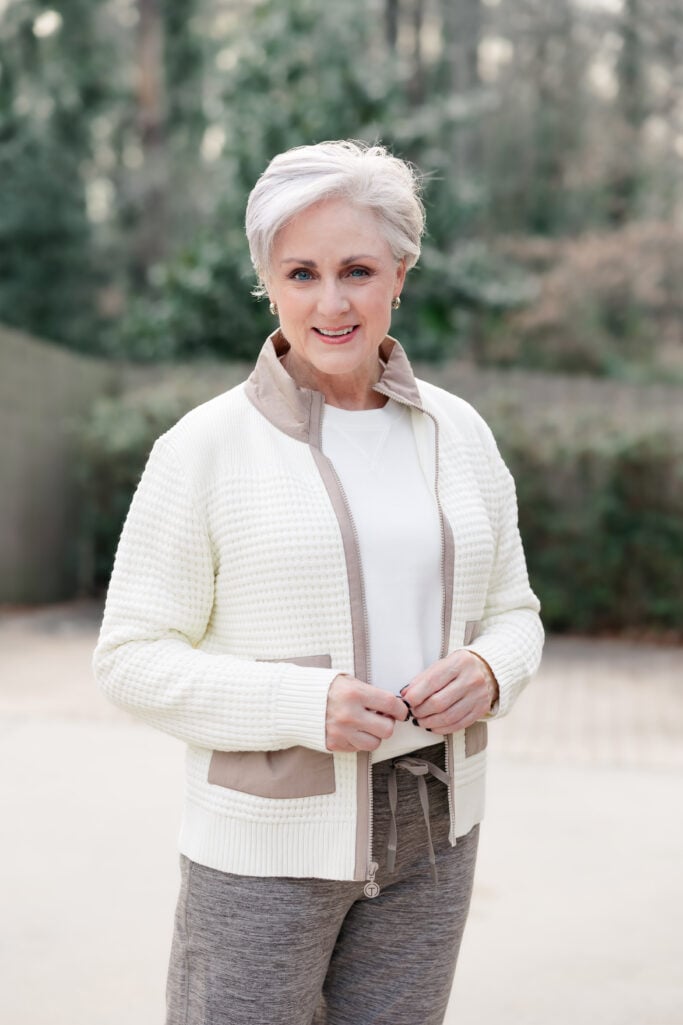 Woman over 50 wearing cream textured sweater jacket with taupe woven pockets and collar over white crewneck top with heather brown joggers