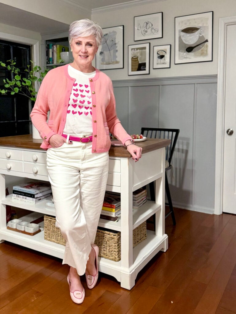 Beth wearing a pink cashmere cardigan with gold buttons over a white hearts graphic tee, white wide-leg crop jeans, a hot pink belt, and pink flats, leaning against the kitchen island