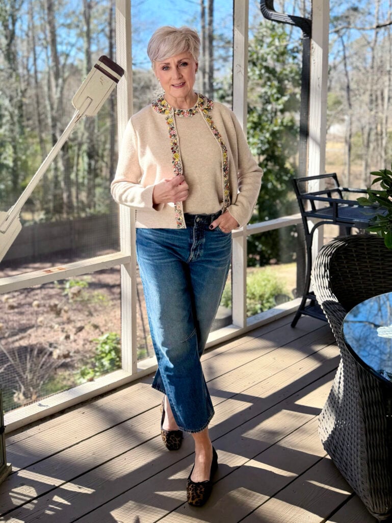 Beth wearing an oatmeal embellished knit cardigan and matching tee with colorful floral beading detail, darted barrel leg jeans, and leopard block heels on the screened porch