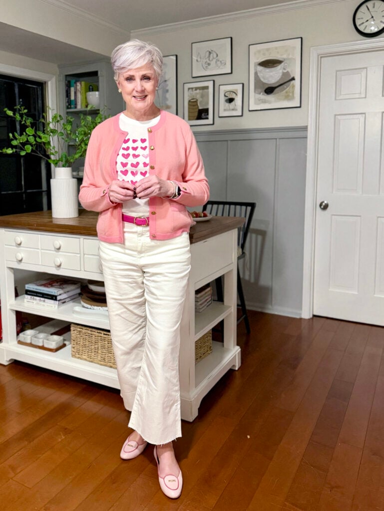 Full-length view of Beth in her pink cardigan, hearts tee, white jeans, and pink flats, standing in front of the kitchen island with art prints on the wall behind her