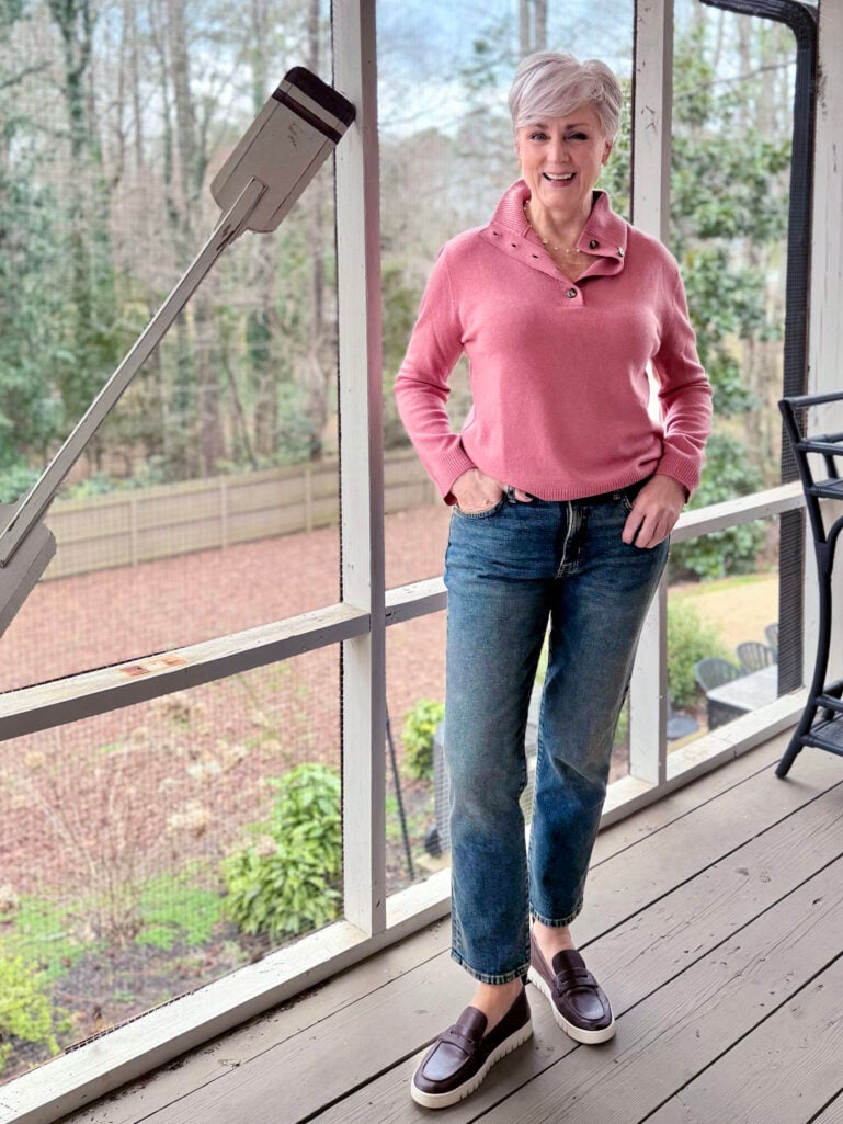 Full-length view of Beth in her pink cashmere sweater, straight jeans, and brown platform penny loafers on the screened porch with winter trees in the background