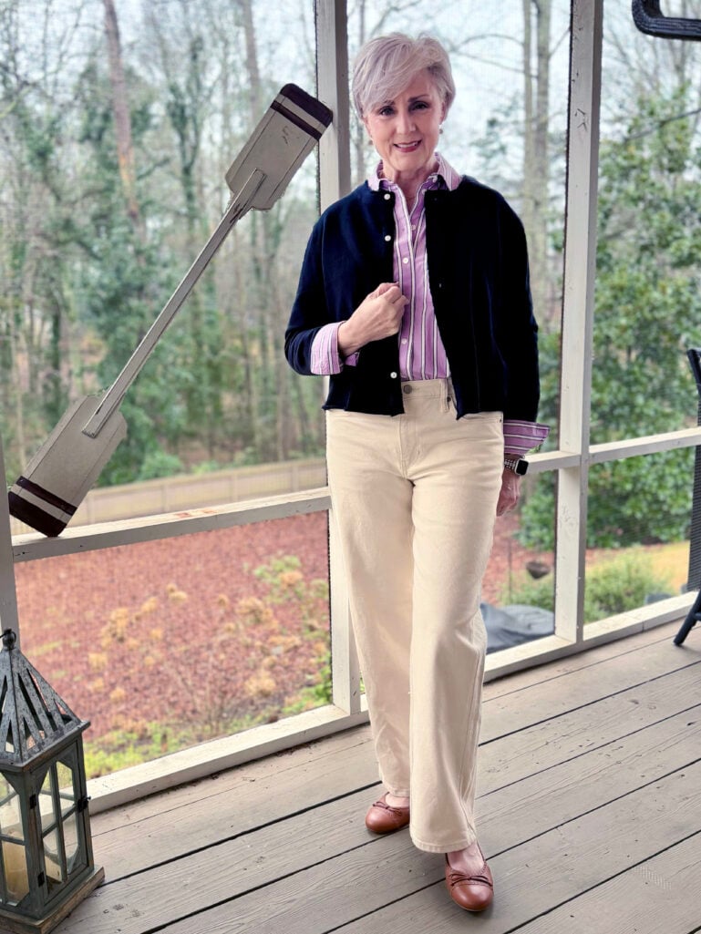 Full-length view of Beth in her navy cardigan and lavender stripes, hand at her collar, winter trees visible through the porch screens