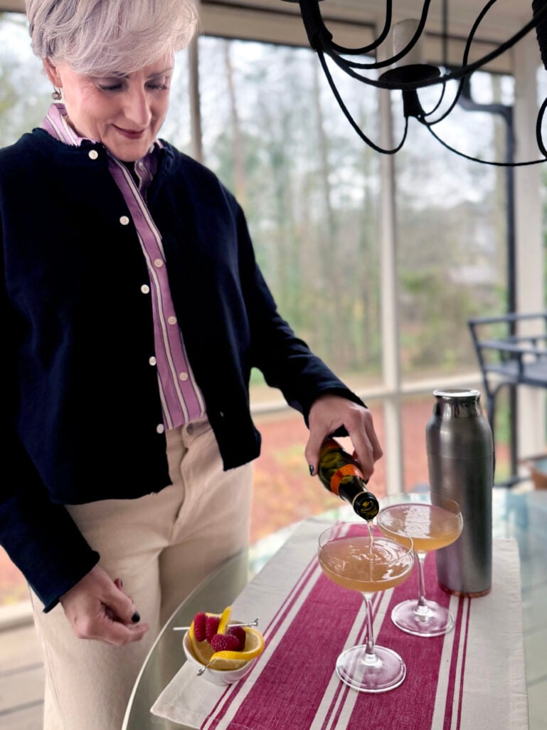 Beth pouring Prosecco into two coupe glasses on the screened porch, a cocktail shaker and bowl of fresh raspberries and lemon beside her