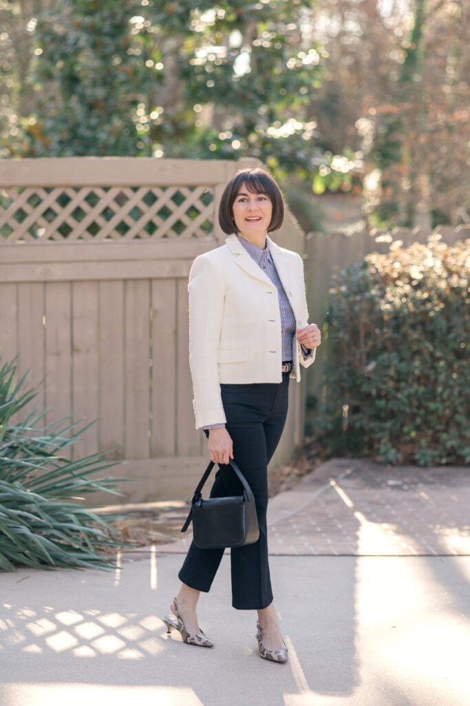 Kelly in ivory bouclé tweed blazer and blue striped button-up shirt with black kick flare jeans, snake print belt, and black wristlet bag — front view of dressed up jeans outfit for petites