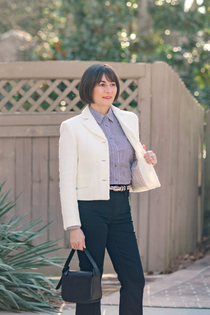 Kelly in ivory bouclé tweed blazer and black striped button-up shirt with black kick flare jeans, snake print belt, and black wristlet bag — front view of dressed up jeans outfit for petites
