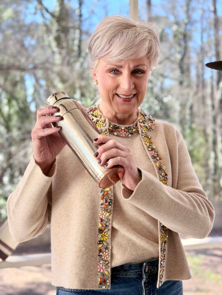 Close-up of Beth shaking a cocktail shaker, smiling, with the colorful floral beading on her embellished cardigan catching the light