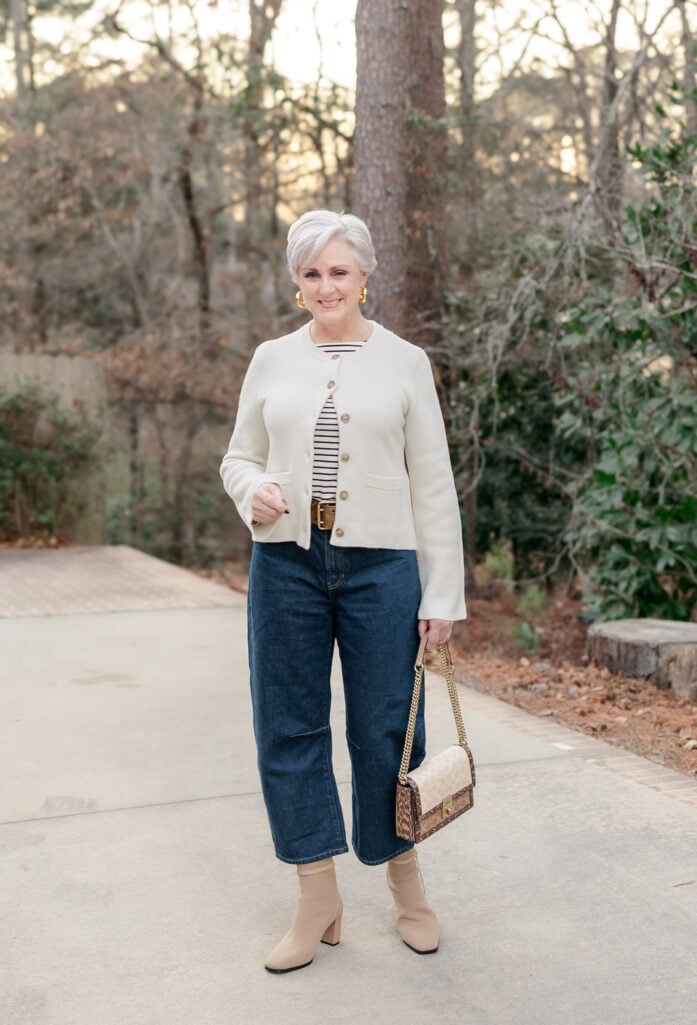 Woman over 50 wearing a classic navy and white Breton striped tee under a cream cashmere structured cardigan with gold buttons, styled with barrel leg jeans, nude square-toe ankle boots, and gold hoop earrings for a timeless French-inspired look.