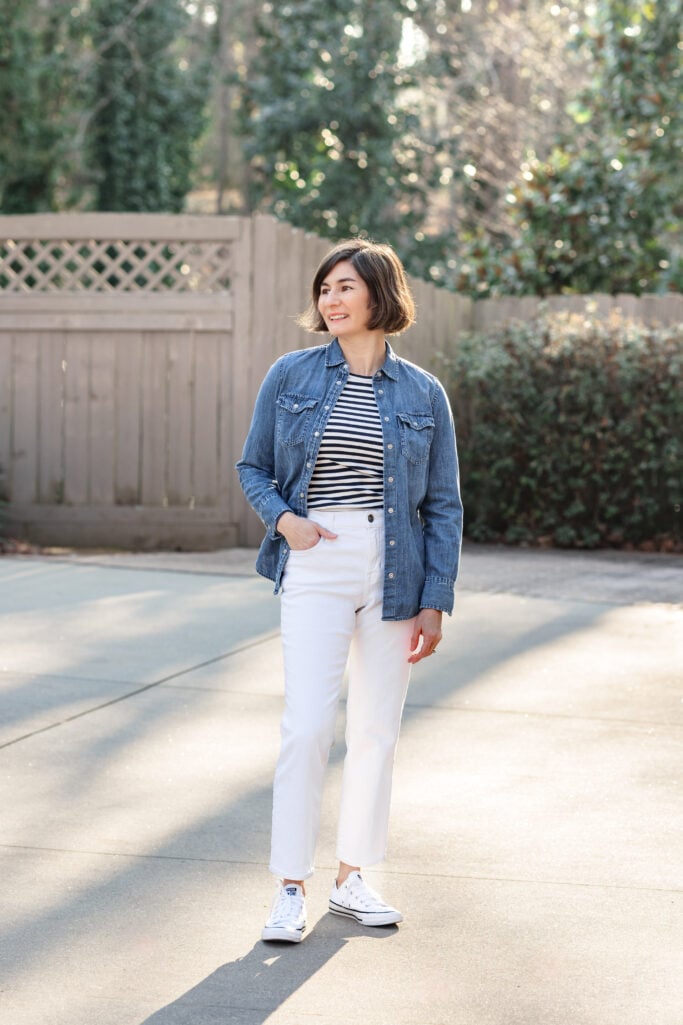 Woman styling a Breton striped tee with an open denim western shirt as a layering piece, white slim jeans, and classic white Converse Chuck Taylor sneakers for a casual weekend outfit idea over 50.