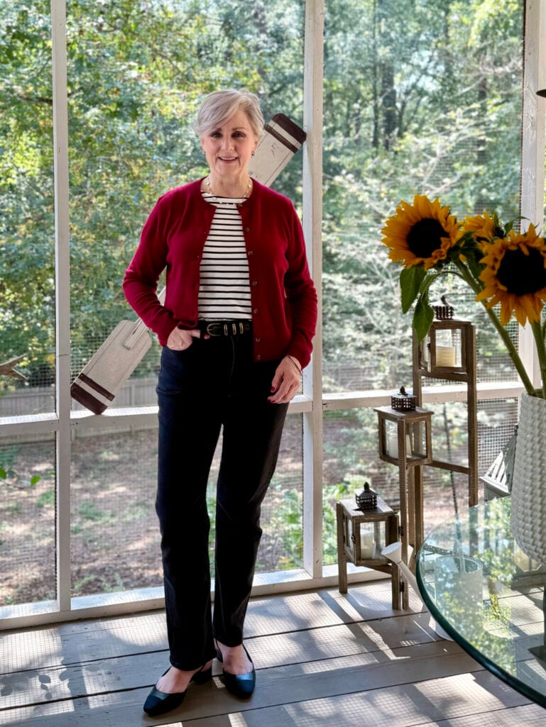 Woman over 50 layering a navy and white Breton striped tee under a burgundy cashmere cardigan with navy trousers, a brown leather belt, and blue cap-toe slingback flats for a polished transitional weather outfit.