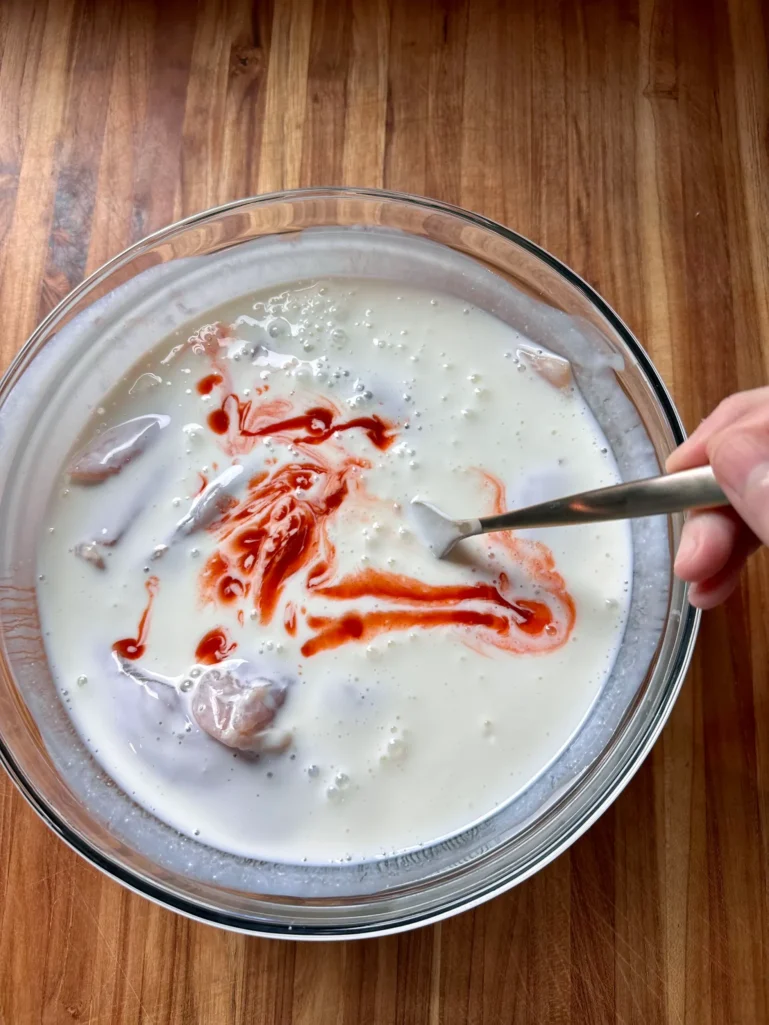 Chicken tenders marinating in buttermilk with hot sauce swirled in, being stirred with a fork