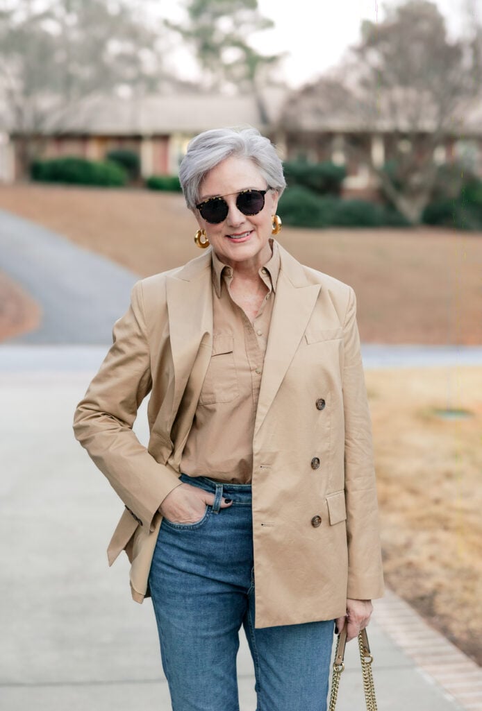 Woman over 50 wearing camel double-breasted blazer over khaki button-down shirt with medium wash jeans, gold chunky hoop earrings, and tortoiseshell sunglasses