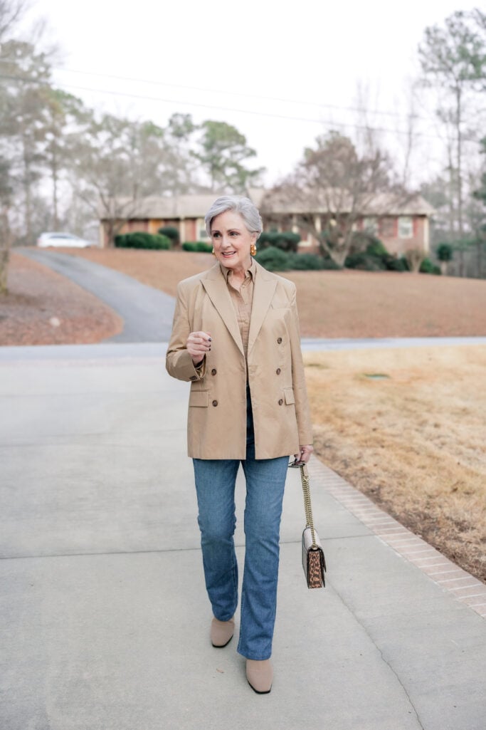 Woman over 50 with silver hair wearing camel blazer and khaki shirt with bootcut jeans, taupe square-toe ankle boots, leopard print crossbody bag, and gold hoop earrings