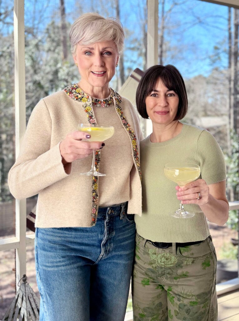 Beth and Kelly holding Lavender Gin Fizz cocktails in coupe glasses, Beth in her embellished cardigan and Kelly in a sage green cashmere tee on the screened porch