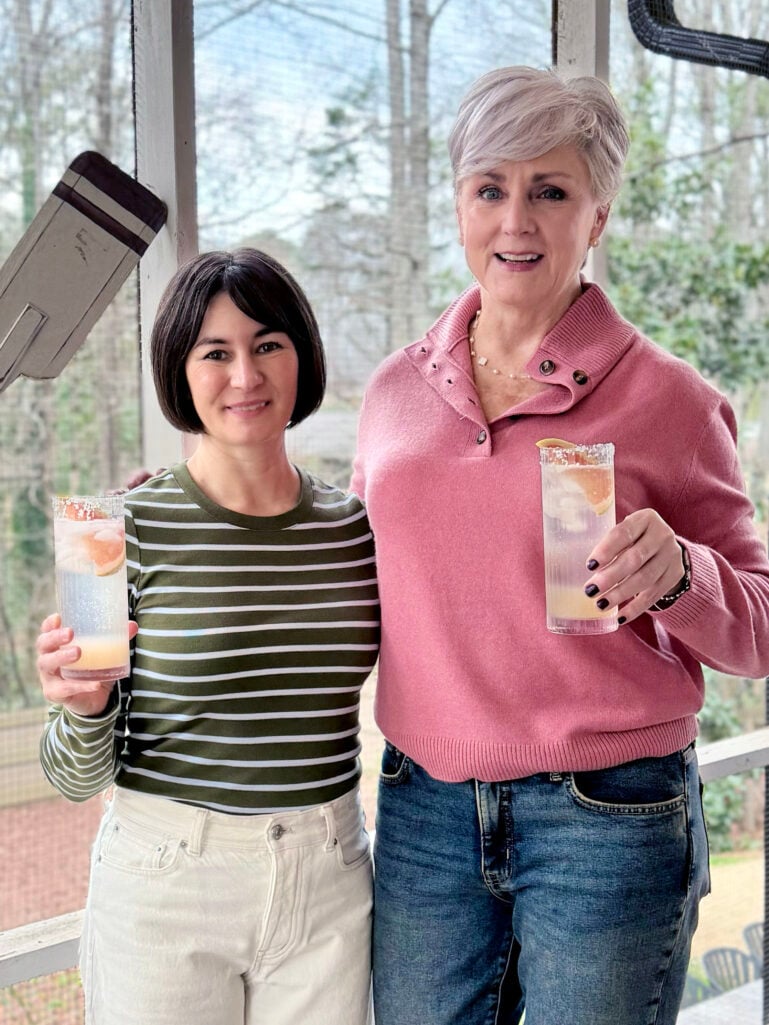Beth and Kelly raising Grapefruit Palomas in tall highball glasses on the screened porch, Beth in her pink sweater and Kelly in an olive striped tee