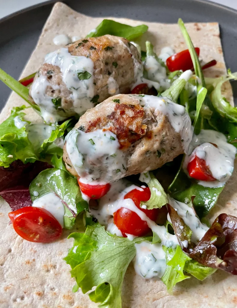 Close-up of broiled chicken kofta drizzled with herbed yogurt sauce on a bed of mixed greens and grape tomatoes over lavash flatbread