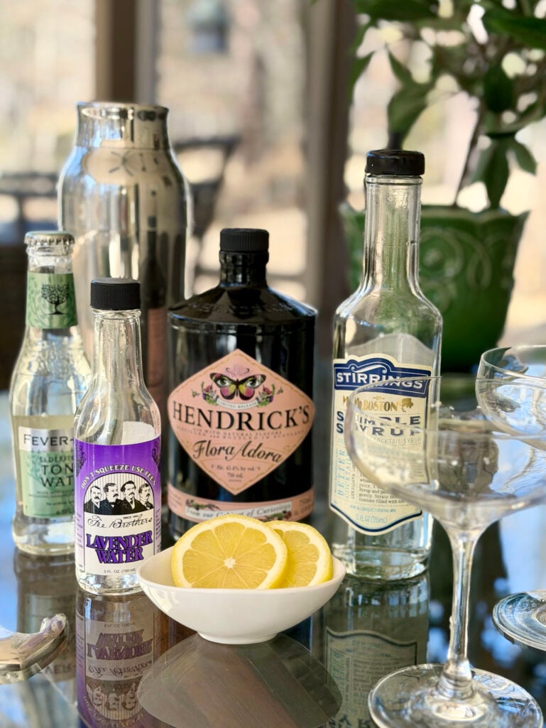 Lavender Gin Fizz ingredients arranged on a glass-topped table: Hendrick's Flora Adora gin, Fever-Tree Elderflower Tonic Water, Fee Brothers Lavender Water, Stirrings Simple Syrup, sliced lemons in a small white bowl, and coupe glasses catching the afternoon light