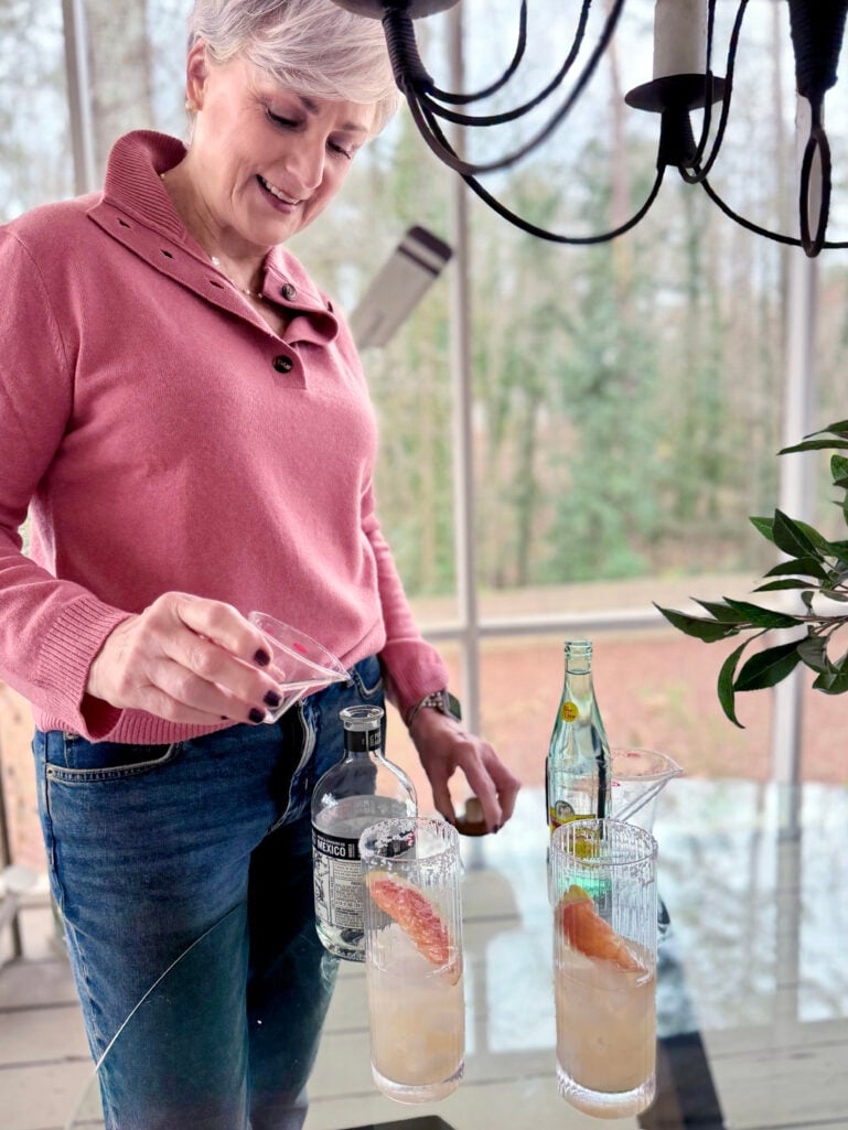 Beth pouring a Grapefruit Paloma from a measuring glass into a tall highball glass garnished with a grapefruit wedge, Espolòn tequila and Topo Chico beside her on the porch
