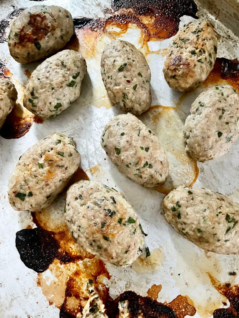 Overhead view of broiled chicken kofta with browned tops and flecks of fresh herbs on a sheet pan