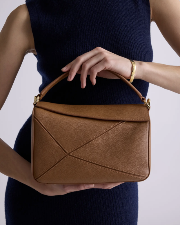 Woman in navy sleeveless knit dress holding structured tan leather top-handle handbag with geometric stitching detail and gold hardware accents.