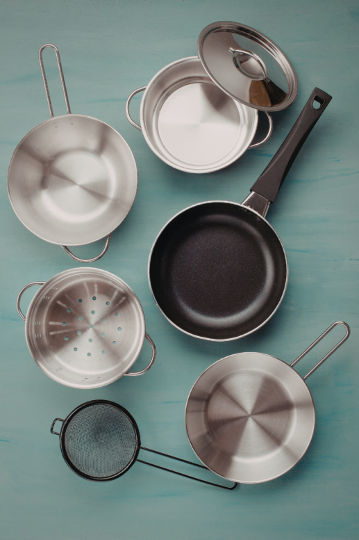 Flat lay of essential cookware including stainless steel saucepans, a nonstick frying pan, a small stockpot with lid, a steamer insert, and a mesh strainer arranged on a teal wooden surface.