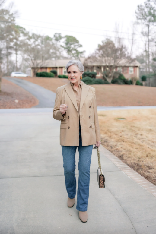 Woman with short gray hair walking down a driveway in a casual, neutral outfit including a tan double-breasted blazer layered over a light brown shirt, straight-leg blue jeans, and beige ankle boots, carrying a small leopard-print shoulder bag with a gold chain strap. Background shows a suburban yard with grass and trees.