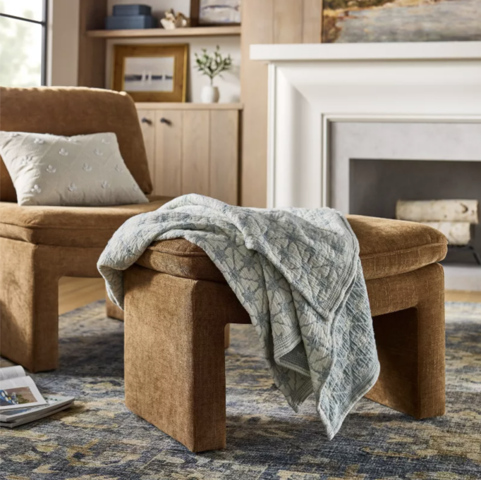 Warm, cozy living room with a caramel velvet lounge chair and matching ottoman, a sage quilted throw draped over the bench, patterned area rug, and white fireplace with light wood built-ins in the background.