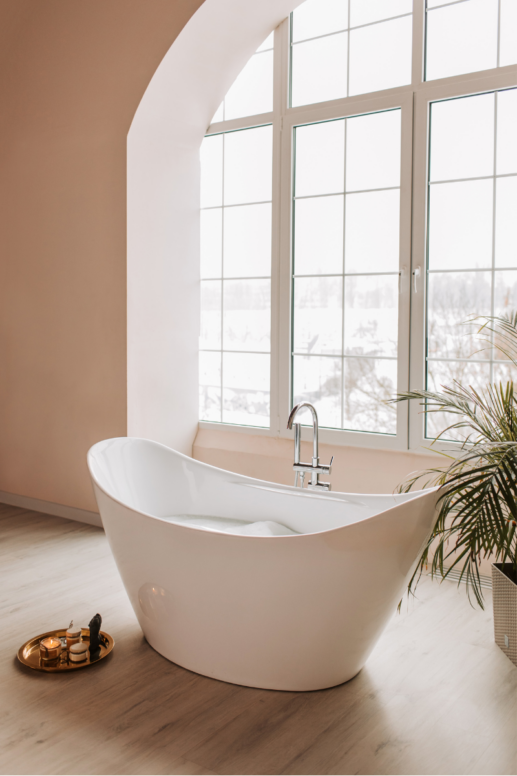 Bright spa-inspired bathroom with freestanding white soaking tub beneath tall arched window, warm peach walls, wood floors, and candle tray for a serene, minimalist look.