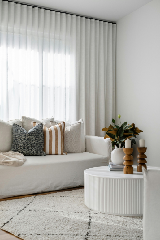 Bright, neutral living room with sheer floor-to-ceiling curtains filtering soft natural light, a white sofa layered with textured throw pillows, a round white coffee table with sculptural wooden candlesticks, and a cozy patterned area rug.