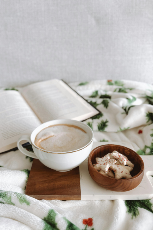 White ceramic mug filled with frothy hot cocoa on a marble and wood serving board, beside a wooden bowl of iced holiday cookies, set on a soft white blanket with green tree pattern and an open book in the background.