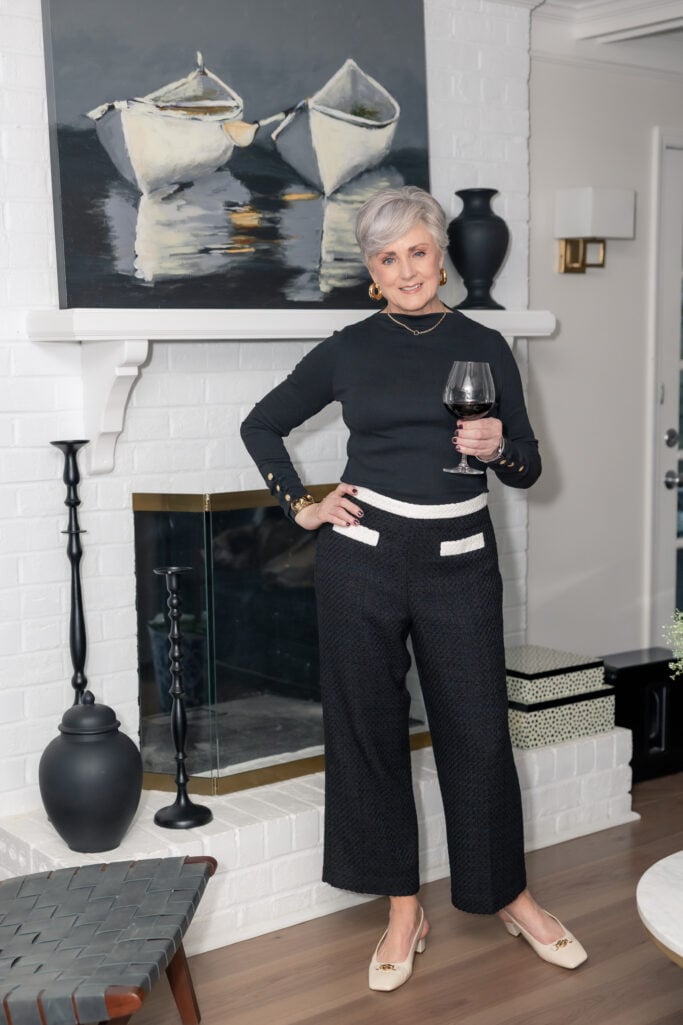 Woman over 50 with silver hair wearing black fitted long-sleeve top with gold button cuff details and black textured wide-leg cropped pants with white contrast waistband and pocket trim, cream block-heel flats with gold hardware, gold hoop earrings and delicate gold necklace