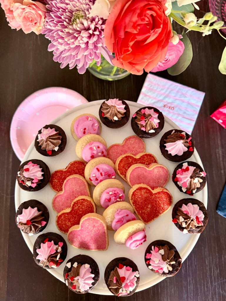 Overhead view of a Valentine's dessert platter with heart-shaped sugar cookies in pink and red frosting and mini chocolate cupcakes topped with pink and chocolate swirled frosting and heart sprinkles, surrounded by pink and red flowers