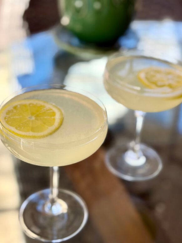 Two Lavender Gin Fizz cocktails in coupe glasses garnished with lemon wheels, soft light on a glass-topped table