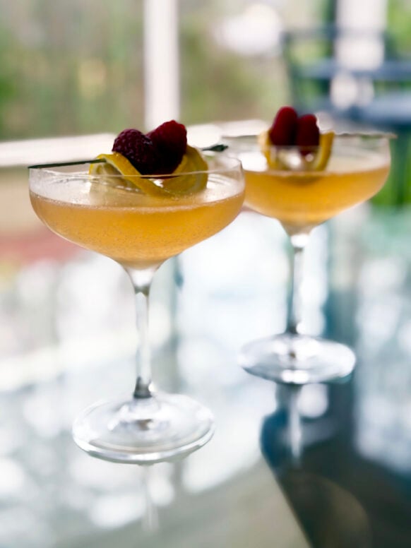 Two Raspberry French 75s in coupe glasses garnished with fresh raspberries and lemon twists, soft porch light reflected on the glass table