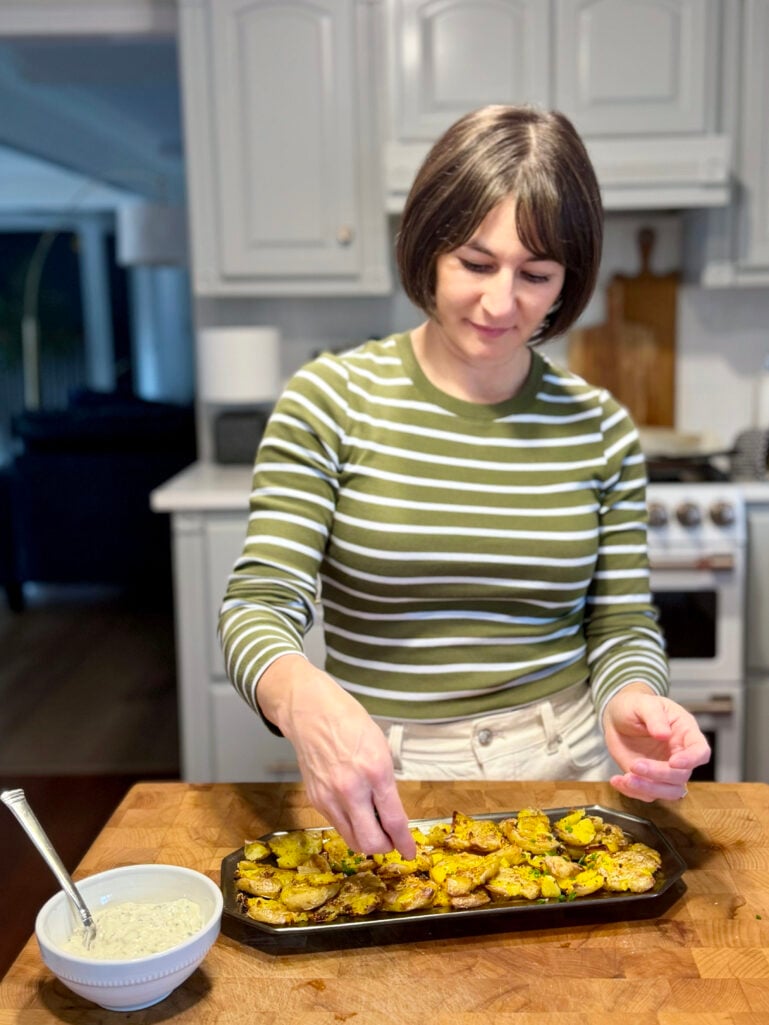 Kelly in her olive striped tee adding finishing touches to a platter of Crispy Smashed Potatoes in the kitchen, a bowl of herbed dipping sauce beside her on the butcher block counter