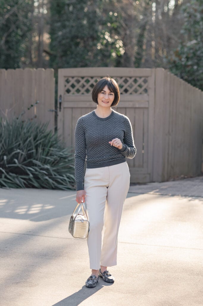 Full-length front view of Kelly in charcoal polka dot sweater, cream ankle pants, plaid loafers, and gold handbag — polished late winter to spring petite outfit