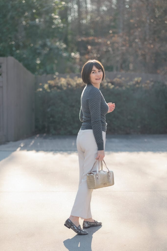 Back view of Kelly walking in charcoal polka dot sweater and cream Quince ankle pants with plaid sequin loafers and gold croc-embossed bag — showing how ankle pants hit at the right length for petites